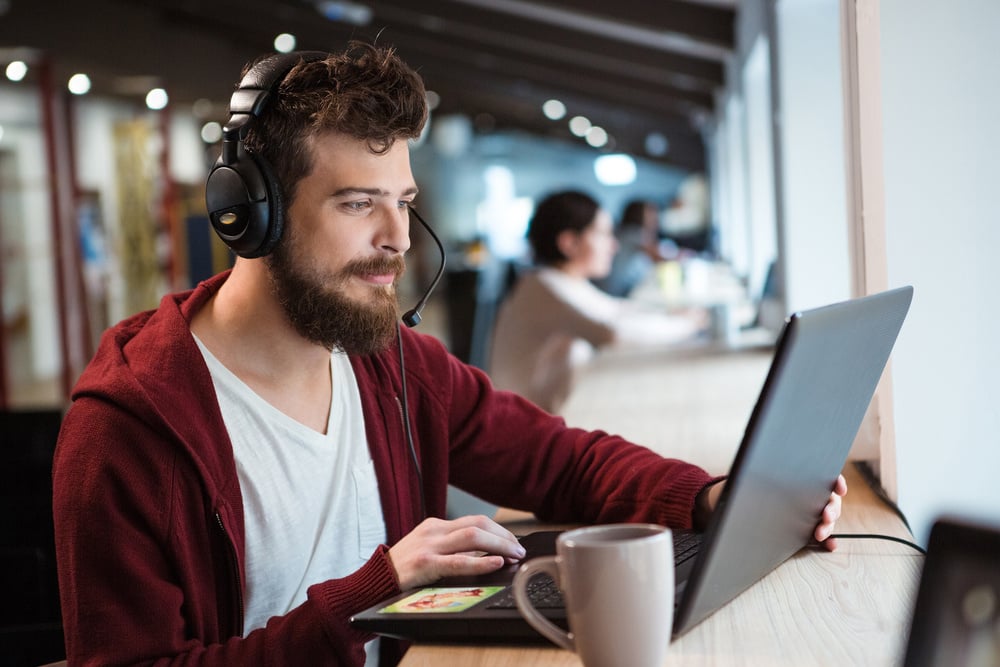 Concentrated handsome male with beard using headset and laptop